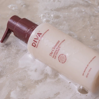 A bottle of DIVA facial cleanser with a maroon pump, lying on a wet surface with bubbles and foam around it. The bottle label says its suitable for dry and sensitive, pH-balanced skin.