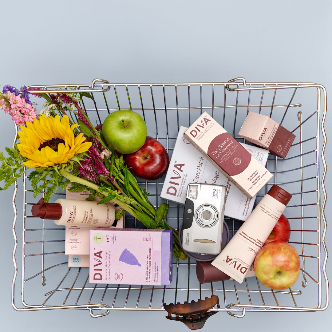 A top-down view of a metal wire shopping cart filled with DIVA skincare products, including bottles, boxes, and a tube, alongside apples, a green apple, flowers, and a small camera, set against a light blue background.