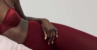 A woman is lying down, wearing a burgundy bra and matching leggings, resting a hand on their thigh against a light pink pillow and neutral background.